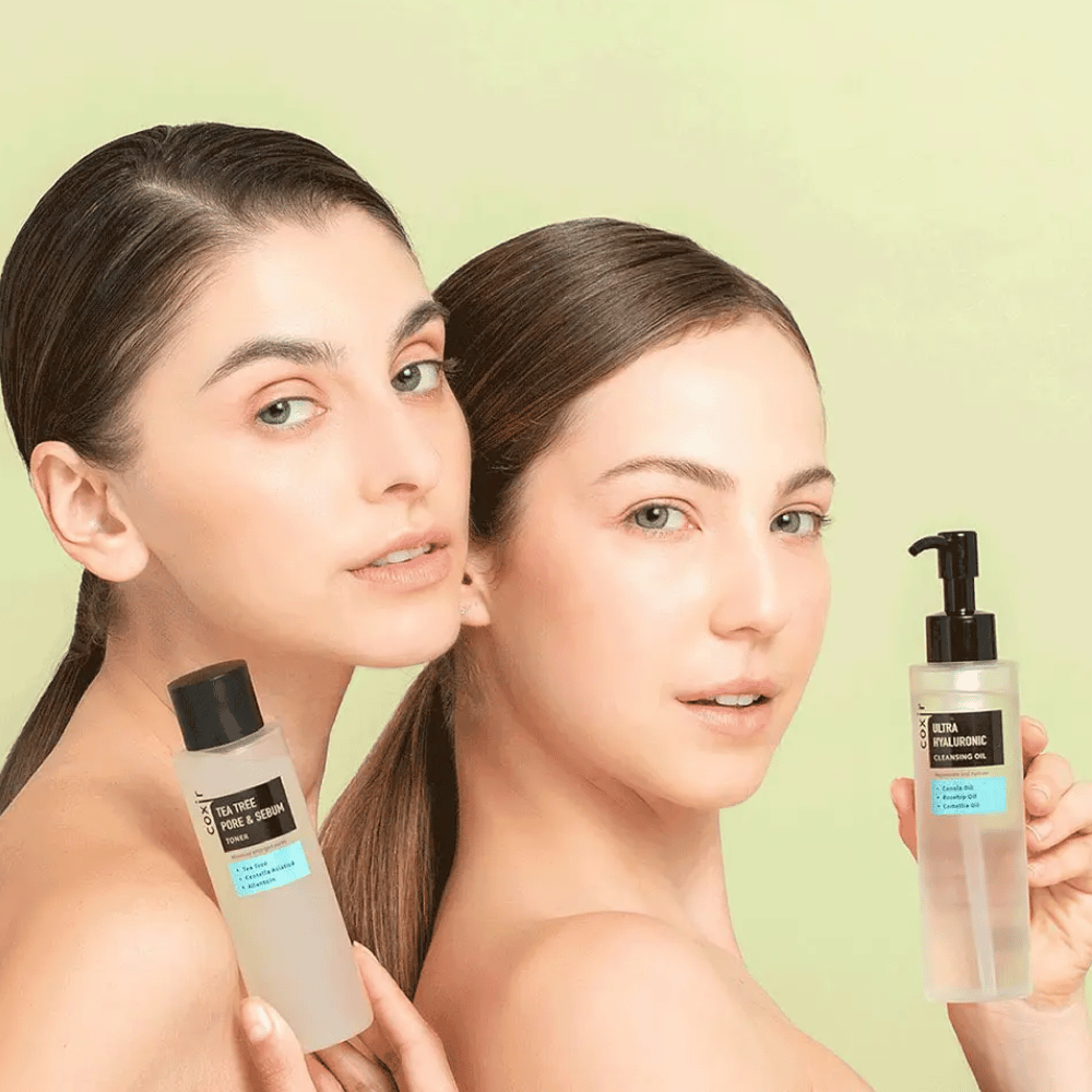 Due donne con le spalle scoperte posano con prodotti per la cura della pelle; una tiene una bottiglia etichettata "TEA TREE PORE & SEBUM", e l'altra un flacone dosatore etichettato "ULTRA HYALURONIC CLEANSING OIL". Stanno davanti a uno sfondo verde chiaro.