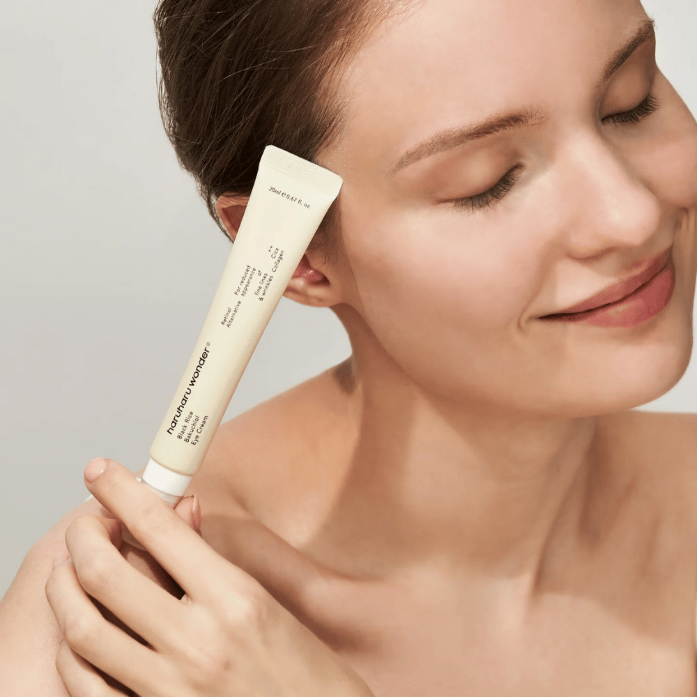 Una persona sorridente con occhi chiusi tiene un tubetto Haruharu Wonder Black Rice Bakuchiol Eye Cream verso la tempia.