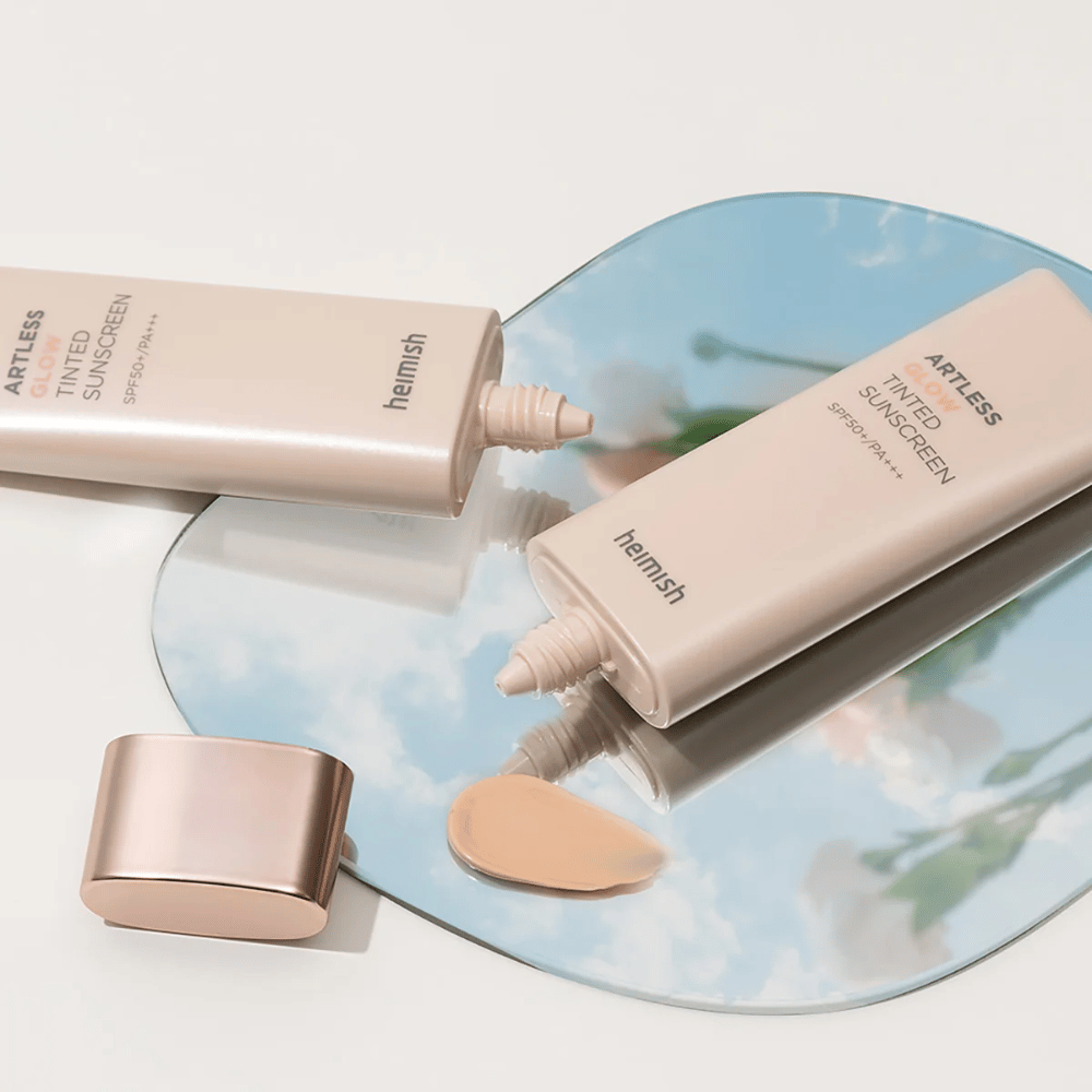 Una bottiglia heimish Artless Glow Tinted Sunscreen SPF50+ PA+++ 40ml e una bottiglia di correttore su uno specchio, che mostra diverse tonalità di pelle.