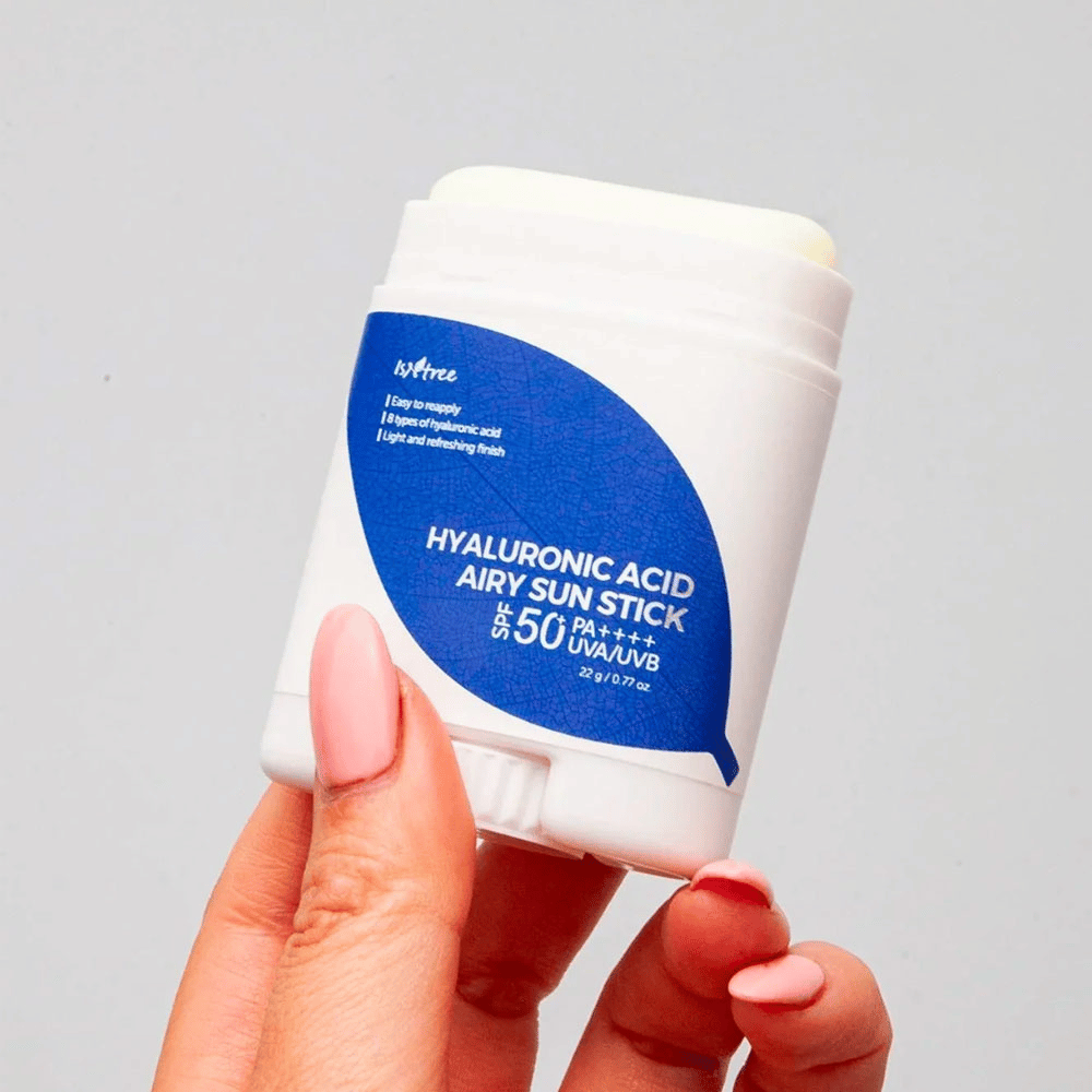 Una mano tiene un bastoncino solare bianco con etichetta blu del marchio Isntree. Il prodotto si chiama "Hyaluronic Acid Airy Sun Stick" e ha un fattore di protezione solare SPF 50+ PA++++.