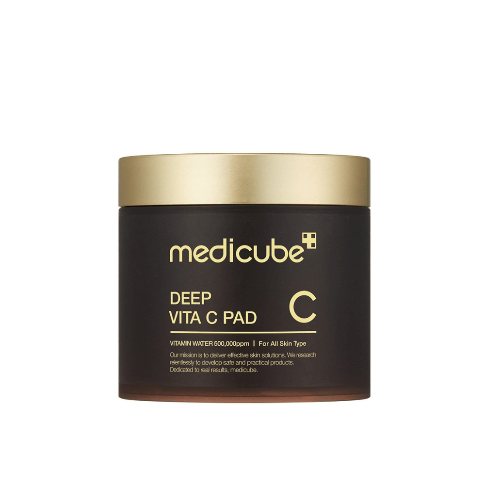 Un barattolo da medicube, etichettato "Deep Vita C Pad", con coperchio dorato e corpo scuro. Il testo sul barattolo indica vitamina C e idoneità per tutti i tipi di pelle. Lo sfondo è neutro.