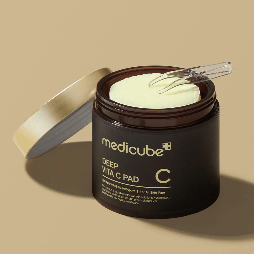 Un barattolo con medicube Deep Vita C Pad. Il barattolo è marrone scuro con testo dorato e un'etichetta bianca invertita con informazioni sulla vitamina C. L'interno del coperchio è dorato, e una pinzetta trasparente si trova sopra i tamponi di cotone giallo chiaro all'interno. Lo sfondo è beige a tinta unita.