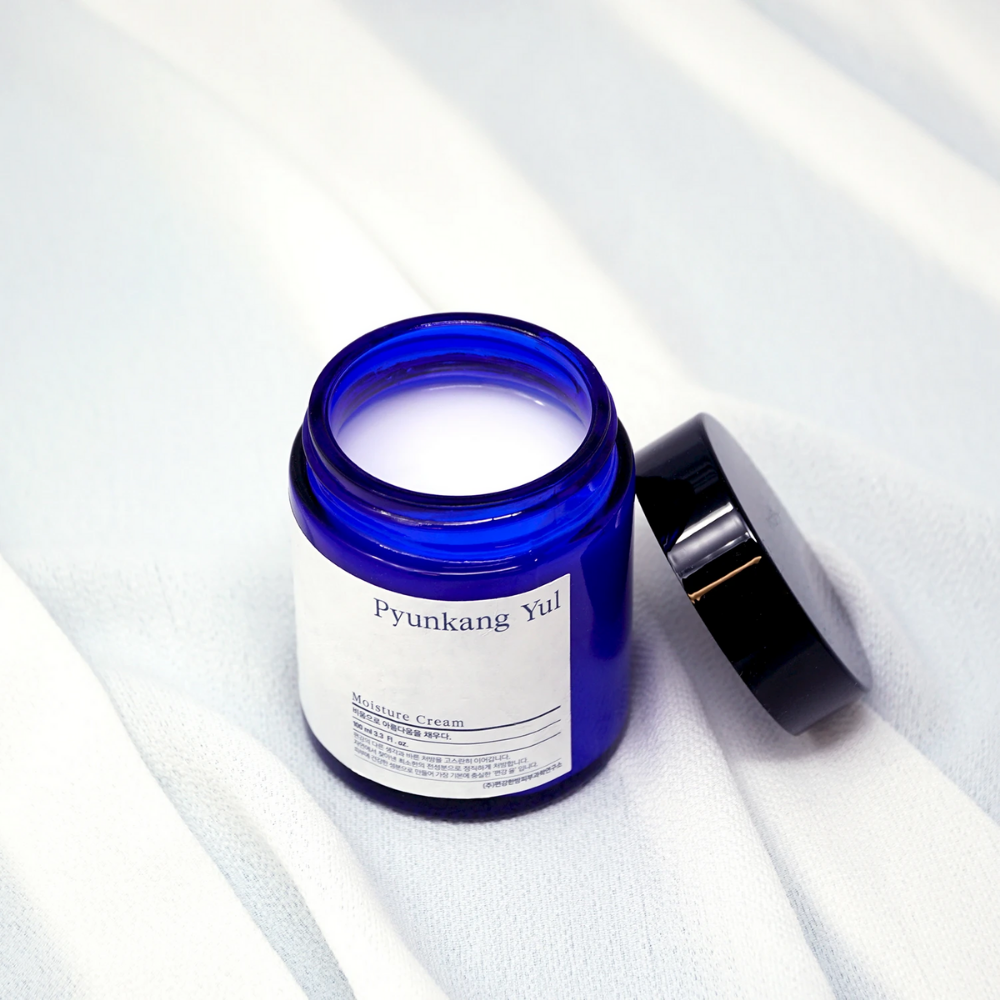 Un barattolo blu con coperchio posizionato su un panno bianco che mostra le proprietà idratanti di Pyunkang Yul's Moisture Cream 100ml.