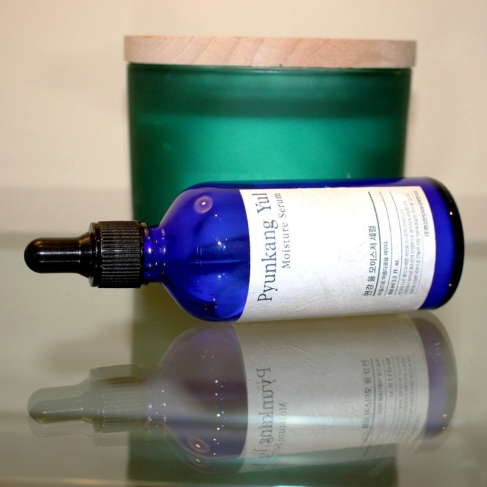 Una bottiglia di Pyunkang Yul Moisture Serum 100ml bilancia i livelli di umidità su un tavolo accanto a una candela.